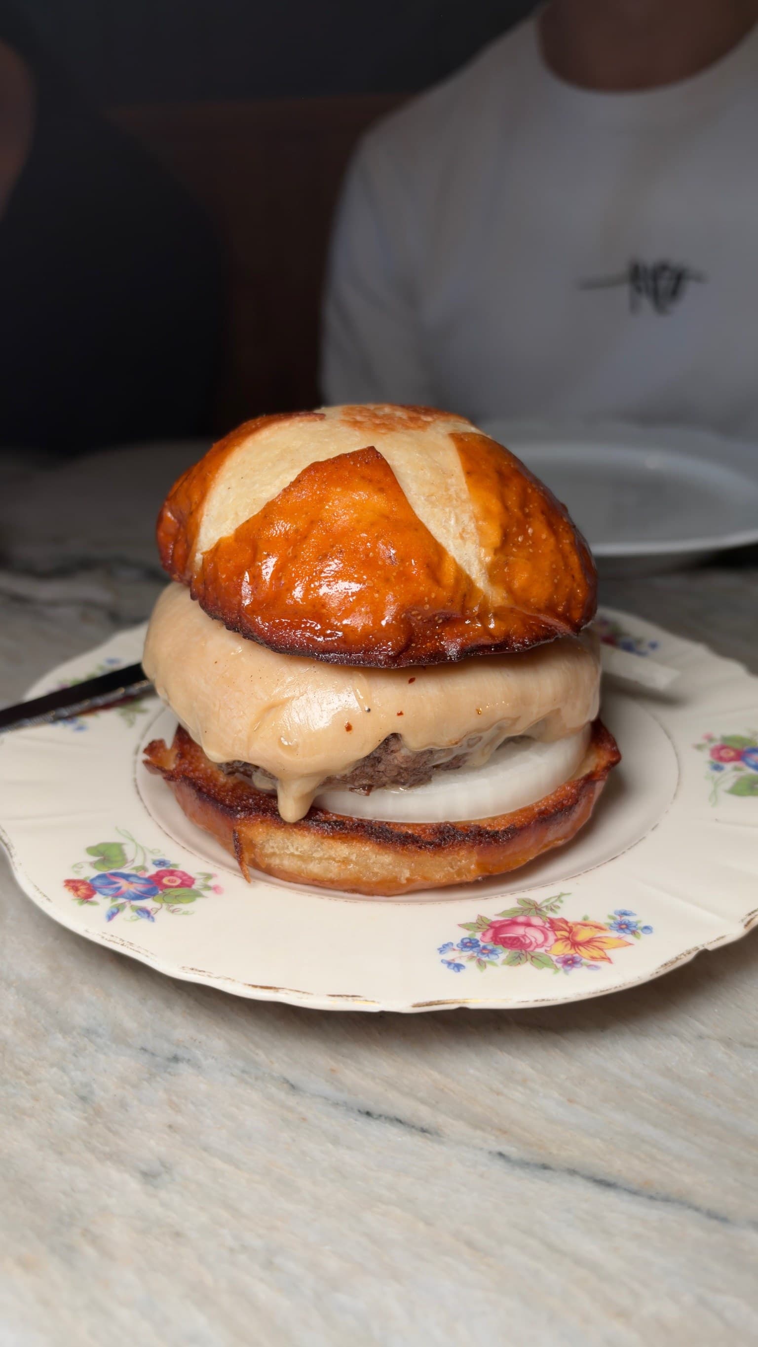 The Welsh Rarebit Cheeseburger at Lord's on a floral china plate: a thick blanket of rarebit over the patty and a slice of raw white onion, under a glossy, scored pretzel bun