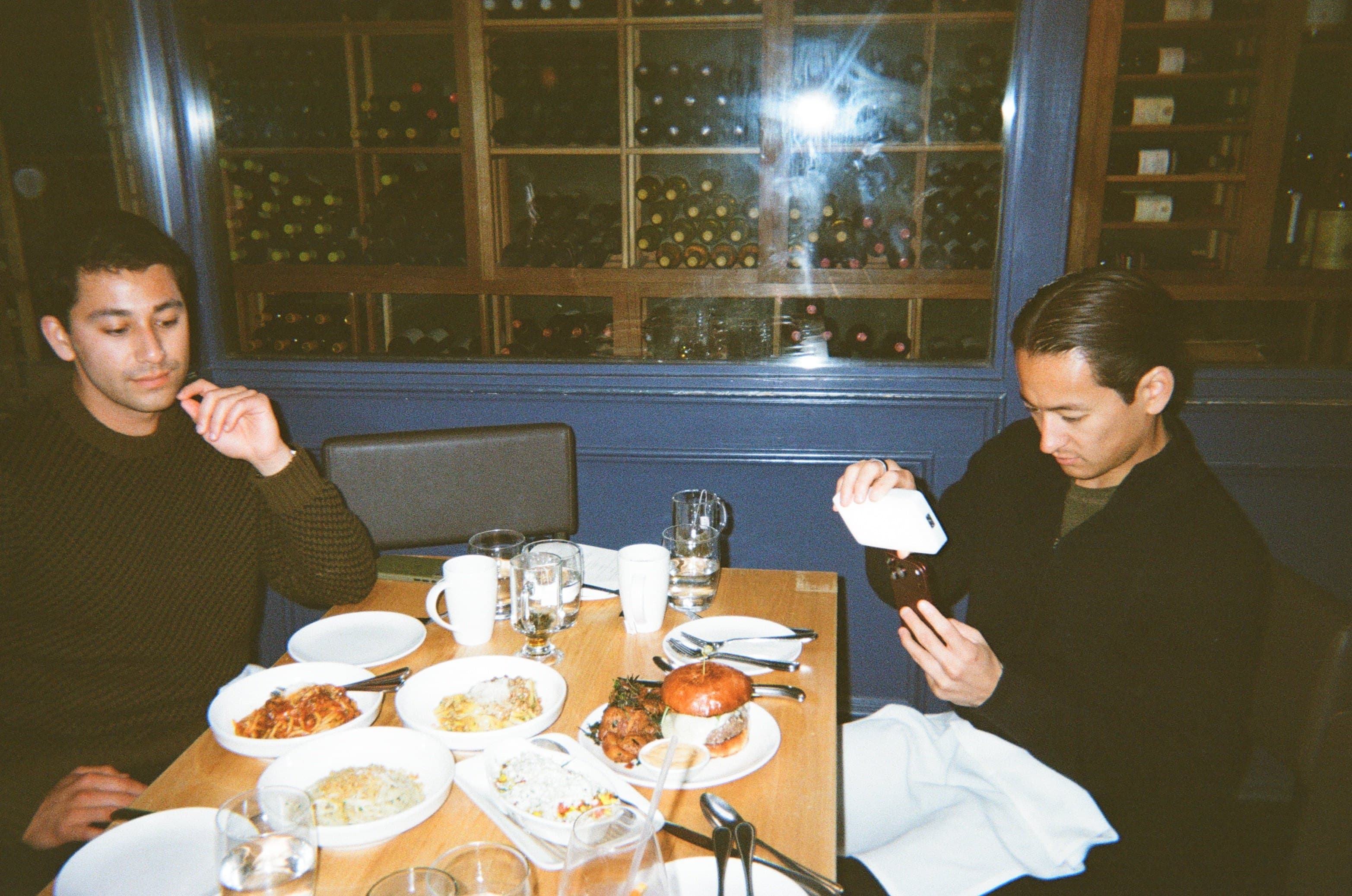 The two brothers behind Brotherly Burgers at a restaurant table in front of a wall of wine bottles, one photographing a burger on his phone, the table set with pasta and sides